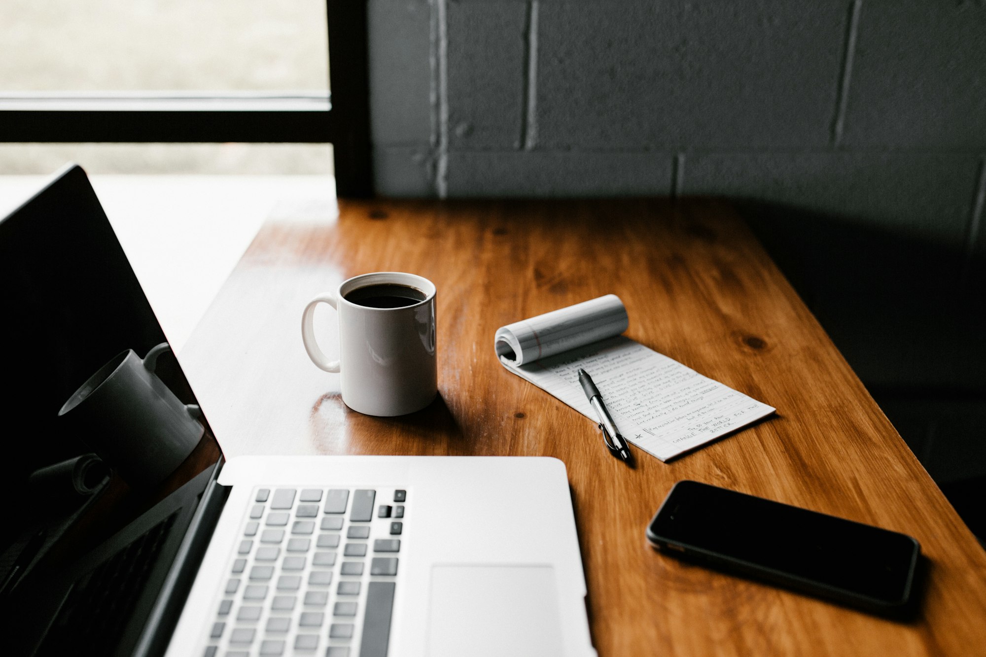 A laptop, notebook, phone and cup of coffee on a wooden desk beside a window.